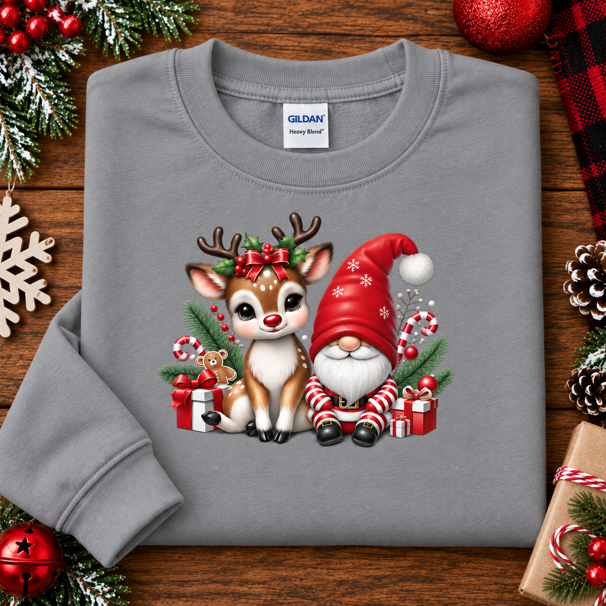 A gray crewneck sweatshirt laid flat on a wooden surface, decorated with Christmas greenery, ornaments, and pinecones. The sweatshirt features a colorful graphic of a cute brown and white reindeer and a red Santa-clad gnome sitting together with candy canes, gifts, and evergreen branches.