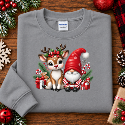 A gray crewneck sweatshirt laid flat on a wooden surface, decorated with Christmas greenery, ornaments, and pinecones. The sweatshirt features a colorful graphic of a cute brown and white reindeer and a red Santa-clad gnome sitting together with candy canes, gifts, and evergreen branches.