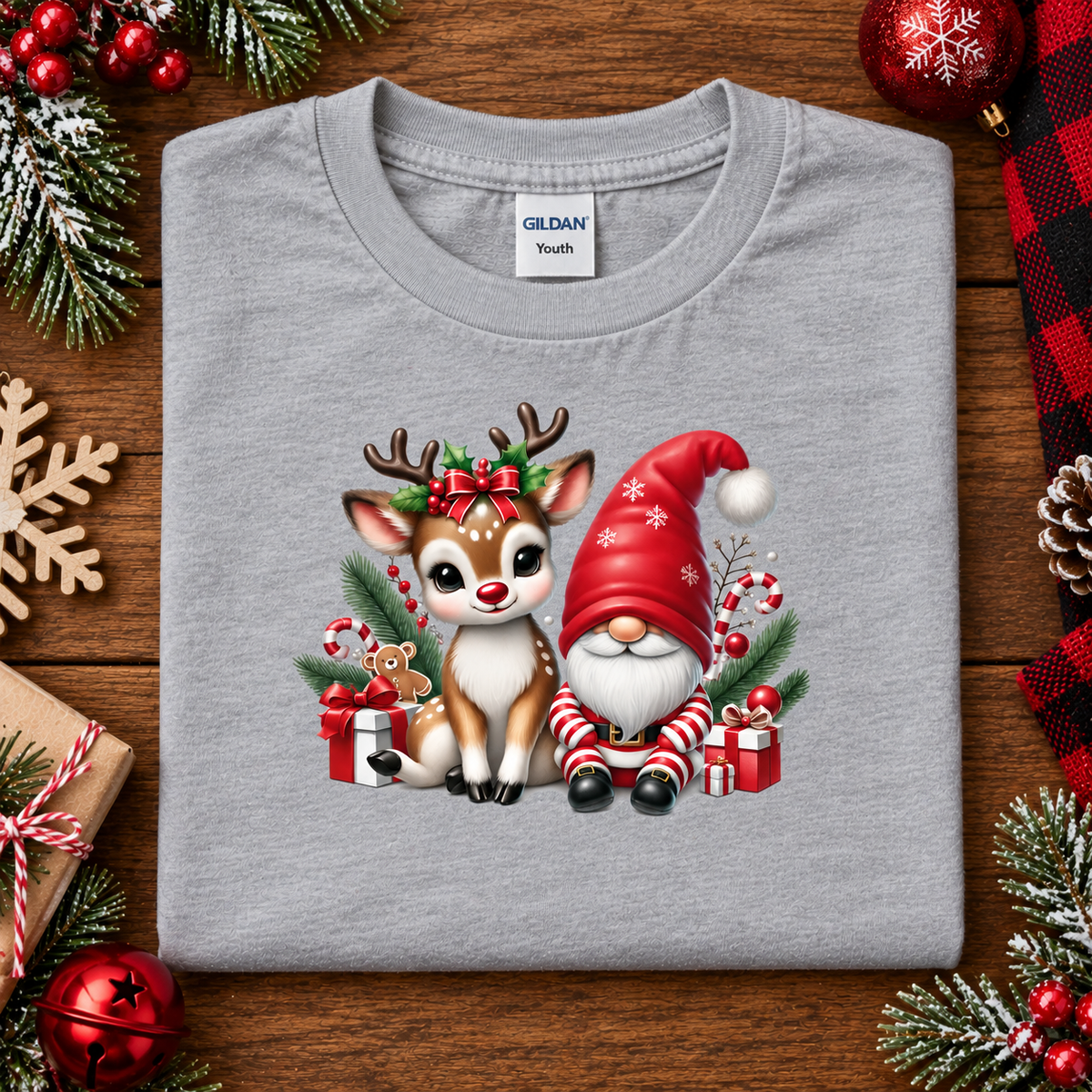 A folded light gray Gildan Youth t-shirt with a festive Christmas graphic of a reindeer and gnome, displayed on a wooden background with holiday decorations.