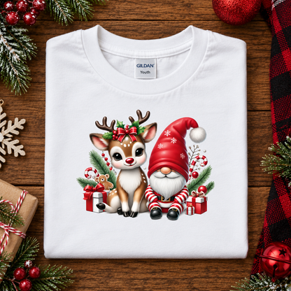 A folded white Gildan Youth t-shirt with a colorful Christmas graphic of a reindeer and a gnome, displayed on a wooden background with holiday decorations.