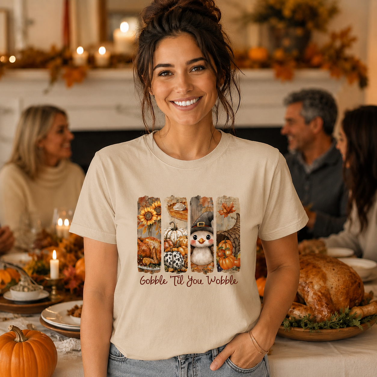 A woman wearing a beige graphic t-shirt with a fall harvest design and the text 'Gobble Till You Wobble' stands in a warmly lit Thanksgiving dining room setting.