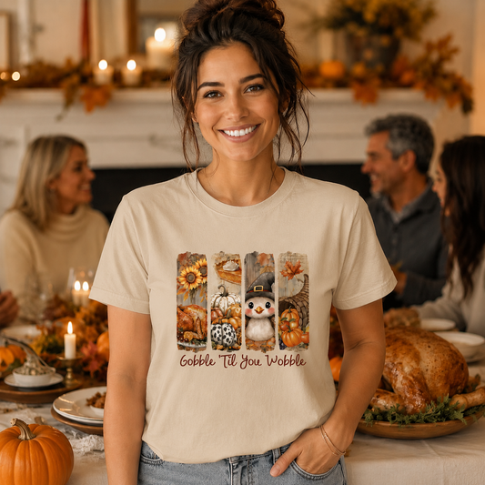 A woman wearing a beige graphic t-shirt with a fall harvest design and the text 'Gobble Till You Wobble' stands in a warmly lit Thanksgiving dining room setting.