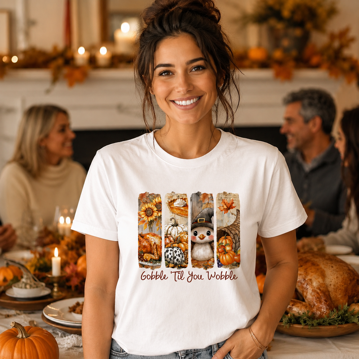 A smiling woman stands at a Thanksgiving dinner table wearing a white graphic t-shirt. The shirt features a vertical collage of autumn-themed illustrations (sunflowers, pumpkins, a snowman, fall leaves) above the text 'Gobble 'til You Wobble' in brown cursive.