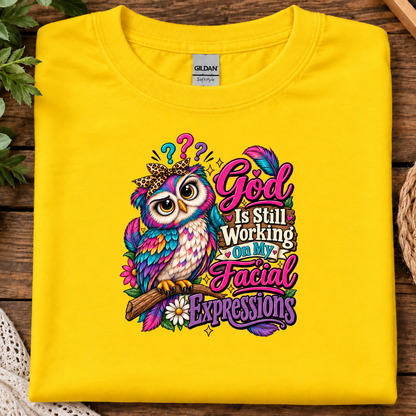 A folded daisy yellow Gildan t-shirt with a colorful graphic print of an owl and the text 'God Is Still Working On My Facial Expressions', displayed on a rustic wooden background.