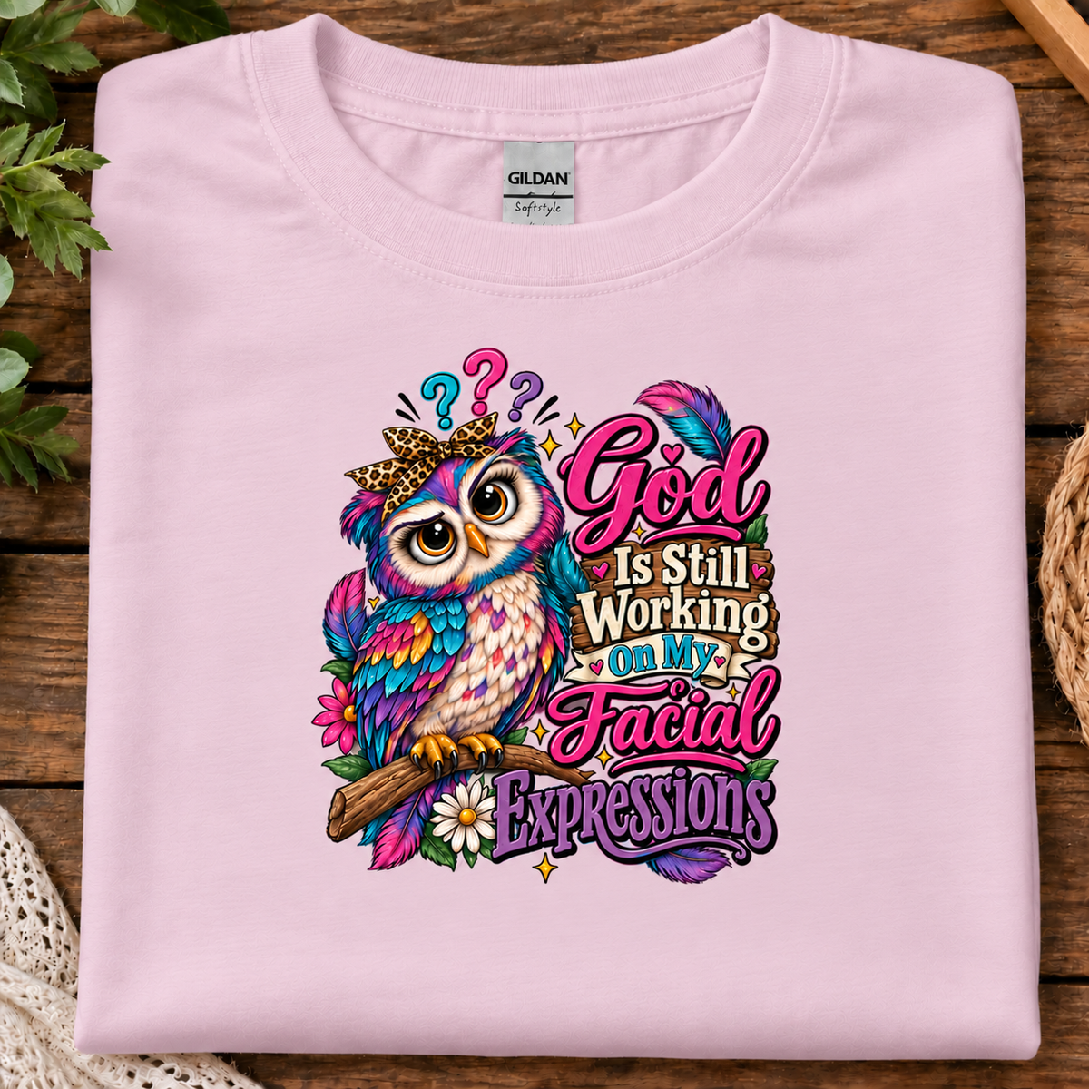 A folded light pink Gildan t-shirt with a colorful graphic of a crowned owl and the text 'God Is Still Working On My Facial Expressions', displayed on a rustic wooden background.