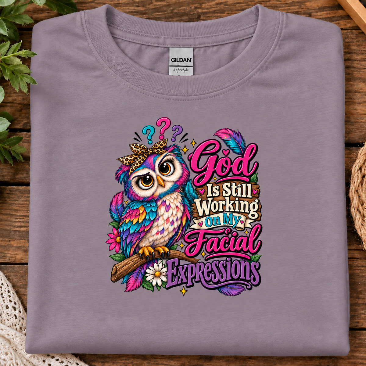 A folded paragon purple Gildan t-shirt with a colorful graphic of a cartoon owl wearing a gold crown and the text 'God Is Still Working On My Facial Expressions', displayed on a rustic wooden background with decorative plants and a basket.