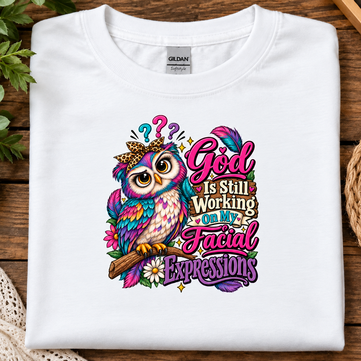 A folded white Gildan t-shirt with a colorful graphic of an owl and the text 'God Is Still Working On My Facial Expressions', displayed on a rustic wooden background with decorative plants and a woven mat.