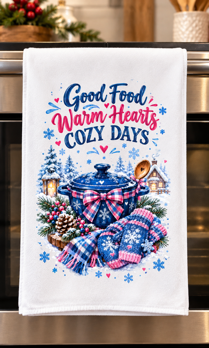 A white kitchen towel hanging on an oven handle, featuring a festive winter design with the text 'Good Food Warm Hearts COZY DAYS' above an illustration of a blue pot, plaid bow, scarf, pinecones, and snowflakes.