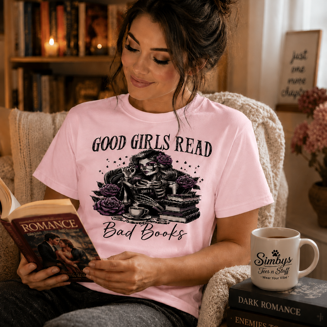 A woman sits in a cozy armchair reading a romance novel, wearing a light pink graphic T-shirt that reads 'GOOD GIRLS READ Bad Books' with a skeleton and books design. A white mug on the chair reads 'Simba's Book & Tea Shop'.