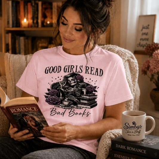 A woman sits in a cozy armchair reading a romance novel, wearing a light pink graphic T-shirt that reads 'GOOD GIRLS READ Bad Books' with a skeleton and books design. A white mug on the chair reads 'Simba's Book & Tea Shop'.