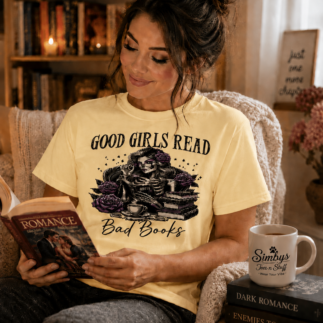A woman sitting in a cozy reading chair, wearing a pale yellow graphic t-shirt that reads 'GOOD GIRLS READ Bad Books' with a skeleton and book illustration. She is holding a romance novel, with a white mug printed with 'Simby's Book & Tea Shop' resting on a stack of books next to her.