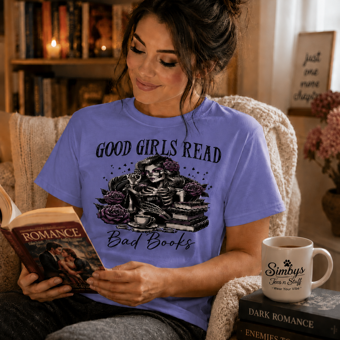 A woman sitting in a cozy armchair, reading a romance novel. She is wearing a lavender graphic t-shirt that reads 'GOOD GIRLS READ Bad Books' with a skeleton and roses illustration.