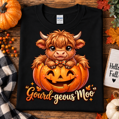 A folded black Gildan Softstyle T-shirt with a cartoon Highland cow sitting in a jack-o'-lantern and the phrase "Gourd-geous Moo" printed below, styled on a wooden surface with autumn props including a pumpkin, fall leaves, berries, and a small sign reading "Hello Fall.".