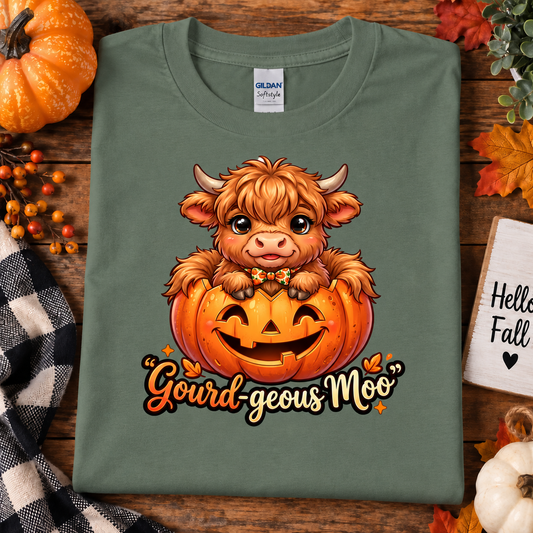 A folded military green Gildan Comfort T-shirt with a cute cartoon Highland cow in a jack-o'-lantern holding a bow tie, with the text 'Gourd-geous Moo' below. The shirt is styled in a flat-lay on a rustic wooden background with autumn decorations including a pumpkin, fall leaves, a checkered cloth, and a small sign that reads 'Hello Fall'.
