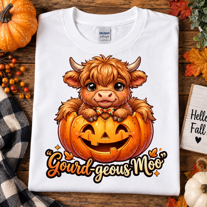 A folded white Gildan Softstyle t-shirt with a cartoon Highland cow in a jack-o'-lantern graphic and the text 'Gourd-geous Moo', styled on a wooden surface with autumn props.