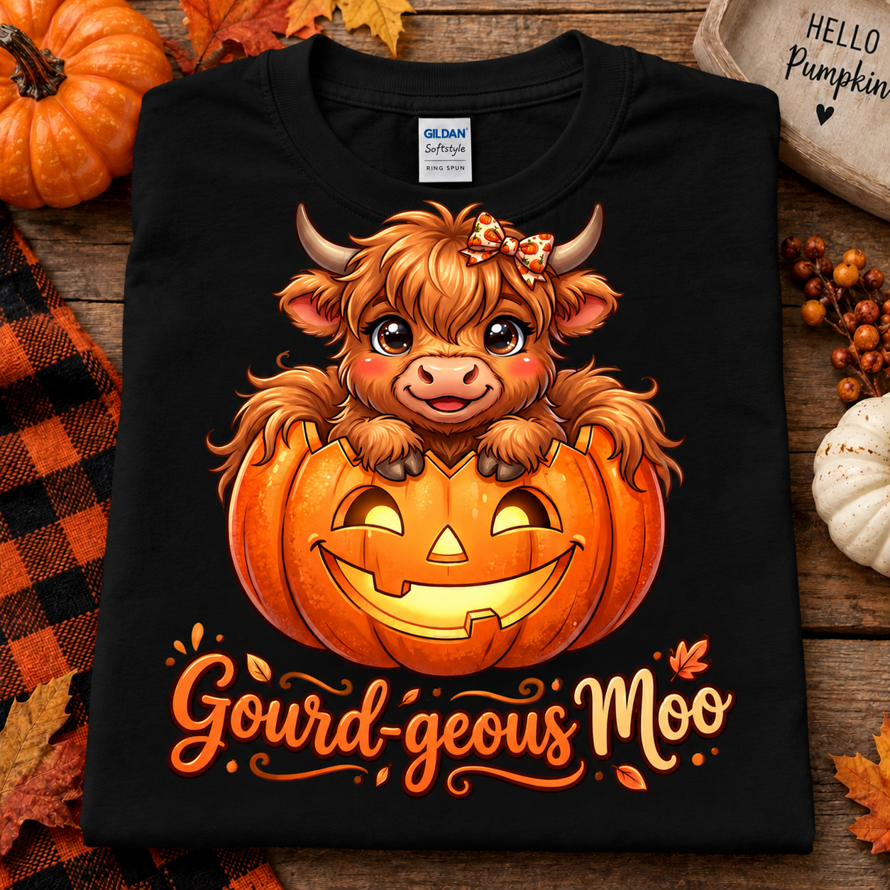 A black Gildan Softstyle t-shirt with a cartoon Scottish Highland cow sitting in a jack-o'-lantern pumpkin, styled on a rustic wooden surface with autumn decorations.