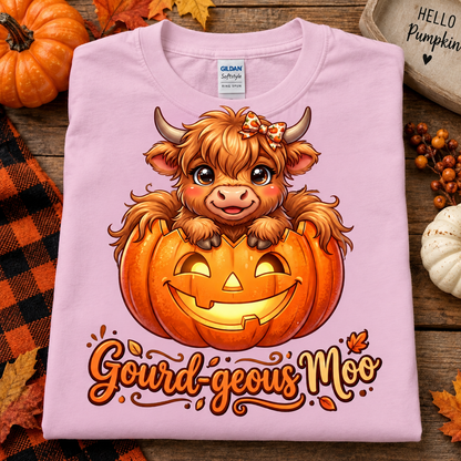 A folded light pink t-shirt with a cartoon illustration of a fluffy orange Scottish highland cow sitting inside a carved jack-o'-lantern pumpkin. Below the illustration is the text 'Gourd-geous Moo' in an orange script font.