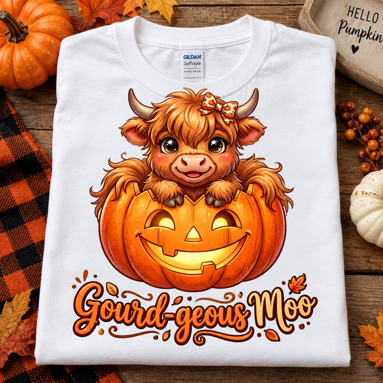 A folded white T-shirt with a colorful graphic of a fluffy Highland cow sitting in a jack-o'-lantern pumpkin, with the phrase 'Gourd-geous Moo' printed below. The shirt is styled on a rustic wooden surface with autumn decorations including pumpkins, leaves, and a small sign.