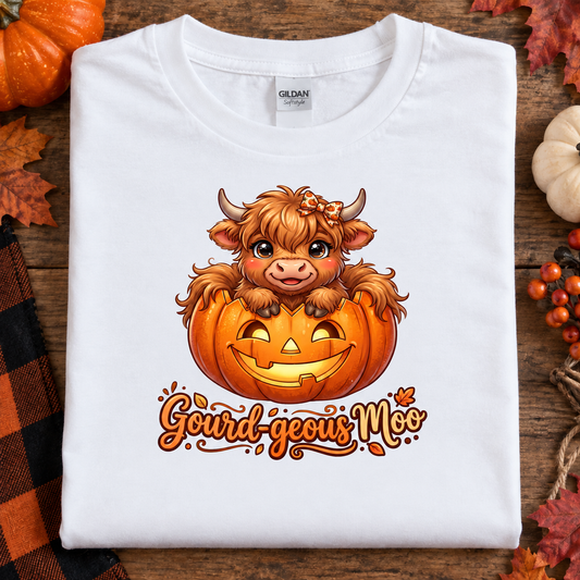 A folded white Gildan t-shirt with a cartoon graphic of a cute Scottish highland cow sitting in a jack-o'-lantern pumpkin, with the text 'Gourd-geous Moo' below. The shirt is displayed on a rustic wooden background decorated with autumn leaves, pumpkins, and a plaid cloth.