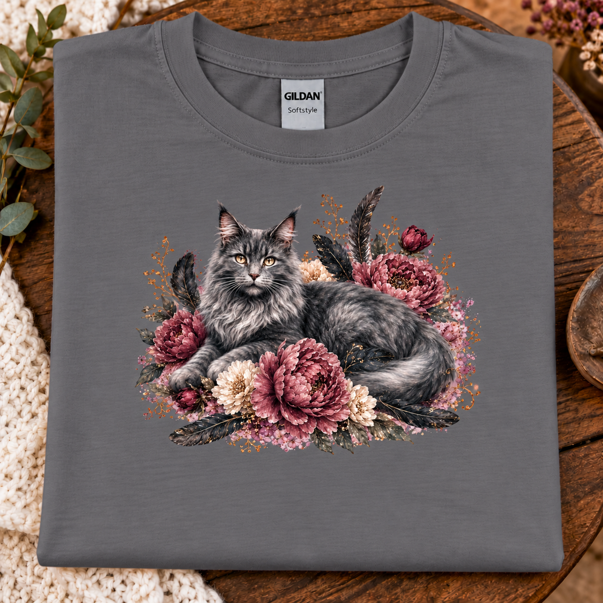 A folded charcola gray Gildan Straightweight t-shirt featuring a detailed graphic of a fluffy gray cat nestled among pink and cream peonies, displayed on a rustic wooden surface with a crochet blanket.