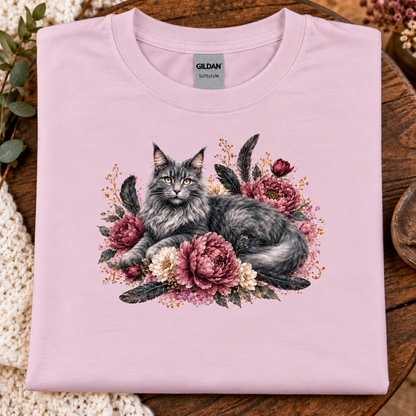 A folded light pink Gildan Softstyle t-shirt featuring a detailed graphic of a fluffy gray cat surrounded by pink and cream peonies, displayed on a rustic wooden surface with a crochet blanket.