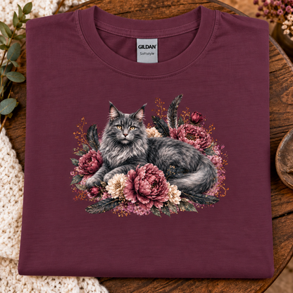 A folded maroon colored Gildan t-shirt in maroon, featuring a detailed graphic of a fluffy gray cat nestled among pink and cream peonies. The shirt is displayed flat on a rustic wooden surface with decorative greenery and a crochet blanket.