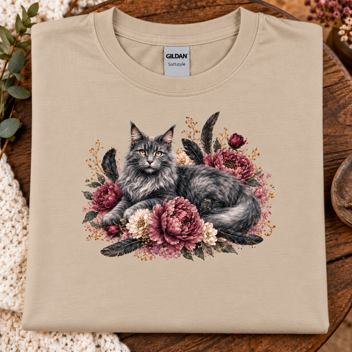 A sand Gildan Softstyle t-shirt laid flat on a rustic wooden surface, featuring a detailed graphic of a fluffy gray cat surrounded by pink and cream peonies with dark feathers.