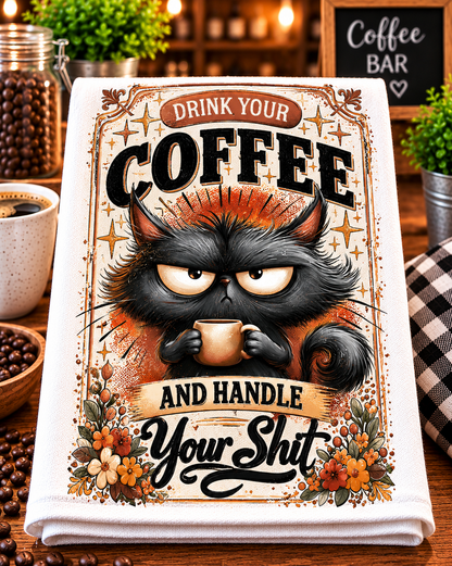 A folded white towel or cloth featuring a humorous coffee-themed graphic print with a grumpy black cat holding a mug and the text 'DRINK YOUR COFFEE AND HANDLE YOUR Shit', displayed on a wooden table in a coffee bar setting.