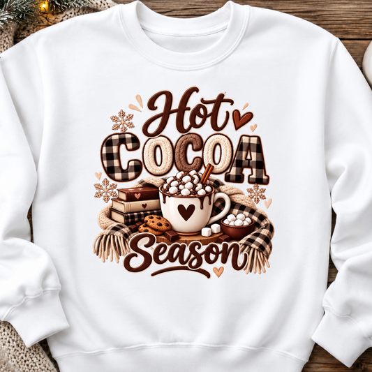 A white crewneck sweatshirt laid flat on a rustic wooden surface, decorated with cozy holiday props like a knit blanket, pinecones, and a small bowl of marshmallows. The front features a large graphic print reading 'Hot Cocoa Season' in brown and beige fonts, surrounding an illustration of a mug filled with marshmallows and cocoa, complete with plaid and textured details.