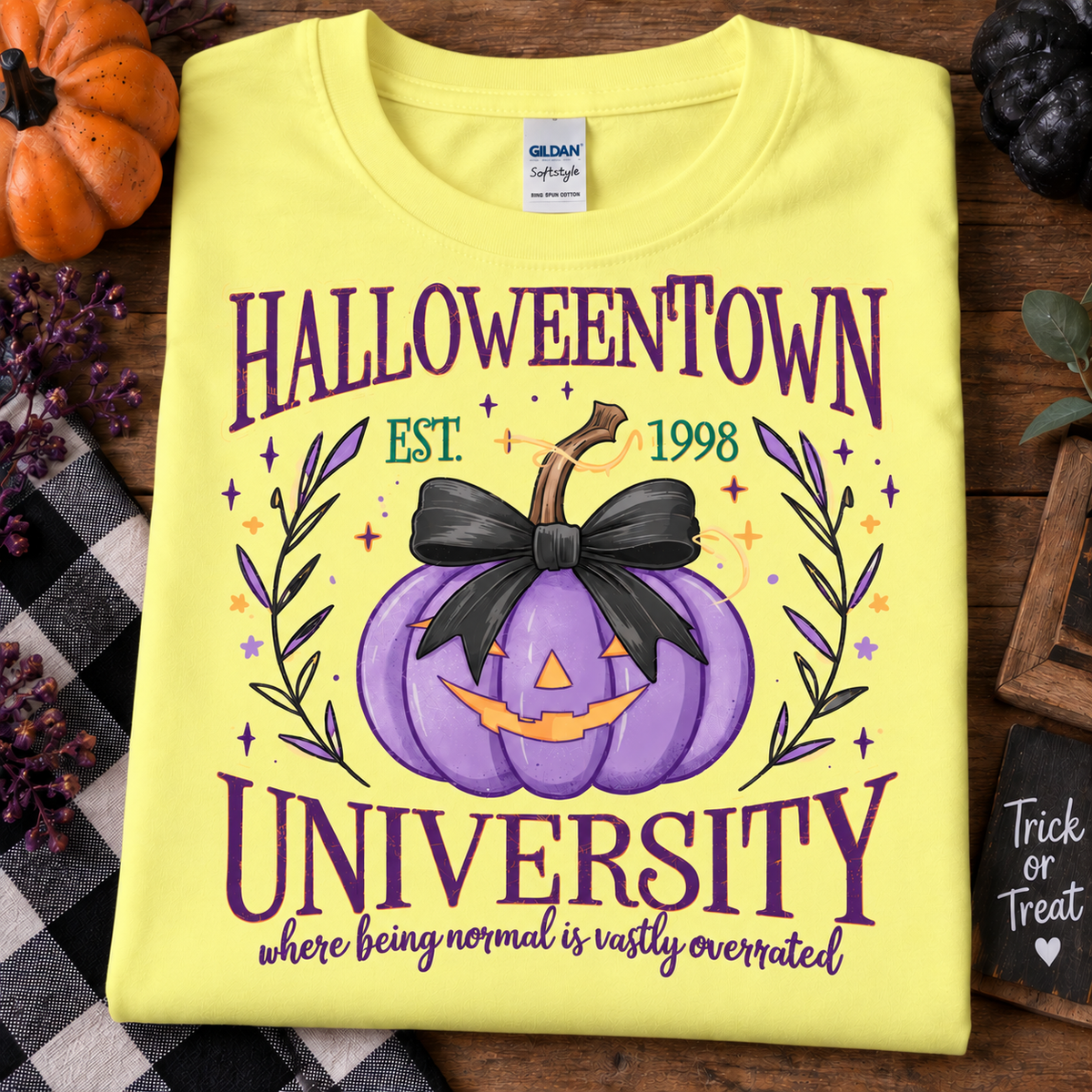 A folded crnsilk yellow Gildan Softstyle T-shirt with a Halloween-themed graphic reading 'HALLOWEENTOWN UNIVERSITY' and 'where being normal is vastly overrated!' around a purple jack-o'-lantern with a black bow. The shirt is styled on a wooden surface with autumn props including a pumpkin, plaid fabric, and a small chalkboard sign.
