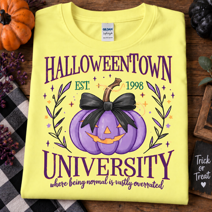 A folded crnsilk yellow Gildan Softstyle T-shirt with a Halloween-themed graphic reading 'HALLOWEENTOWN UNIVERSITY' and 'where being normal is vastly overrated!' around a purple jack-o'-lantern with a black bow. The shirt is styled on a wooden surface with autumn props including a pumpkin, plaid fabric, and a small chalkboard sign.