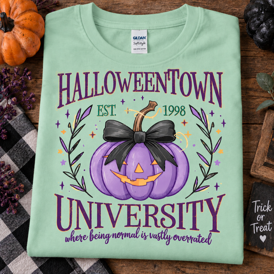 A mint green Gildan Softstyle T-shirt laid flat on a rustic wooden surface, featuring a Halloween-themed graphic that reads 'HALLOWEENTOWN UNIVERSITY' with a purple jack-o'-lantern, black bow, and decorative text. The shirt is styled with autumn props including a small orange pumpkin, a plaid blanket, and a chalkboard.