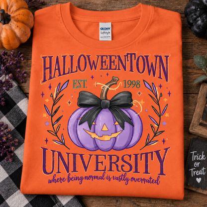 An orange Gildan Softstyle T-shirt laid flat on a rustic wooden surface, featuring a Halloween-themed graphic that reads 'HALLOWEENTOWN UNIVERSITY' with a purple jack-o'-lantern and black bow. Decorative pumpkins, a plaid cloth, and fall props surround the shirt.