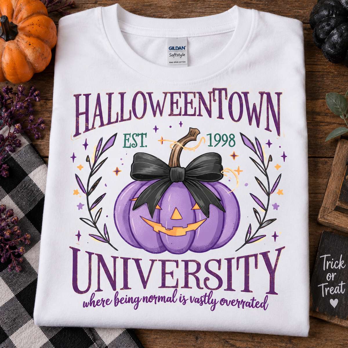 A folded white Gildan Softstyle T-shirt with a Halloween-themed graphic print reading 'HALLOWEENTOWN UNIVERSITY' and 'where being normal is vastly overrated,' featuring a purple pumpkin with a black bow and orange details, styled on a wooden surface with autumn props.