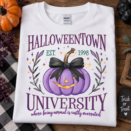 A folded white Gildan Softstyle T-shirt with a Halloween-themed graphic print reading 'HALLOWEENTOWN UNIVERSITY' and 'where being normal is vastly overrated,' featuring a purple pumpkin with a black bow and orange details, styled on a wooden surface with autumn props.
