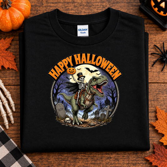 A folded black Gildan Youth t-shirt with a 'Happy Halloween' graphic of a skeleton riding a T-Rex dinosaur, displayed on a wooden surface with autumn props.