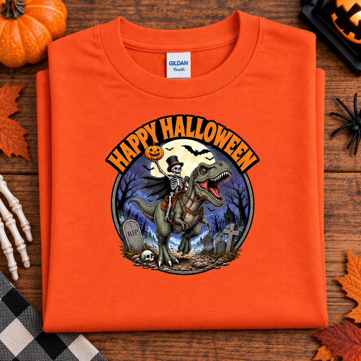 An orange Gildan Tees folded t-shirt with a 'Happy Halloween' graphic featuring a skeleton riding a T-Rex dinosaur in a graveyard, displayed on a wooden surface with seasonal props.