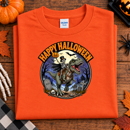 An orange Gildan Tees folded t-shirt with a 'Happy Halloween' graphic featuring a skeleton riding a T-Rex dinosaur in a graveyard, displayed on a wooden surface with seasonal props.