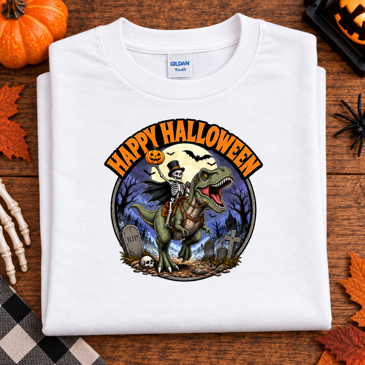 A folded white Gildan Softstyle T-shirt with a colorful Halloween graphic showing a skeleton riding a green T-rex dinosaur in a graveyard, with the text 'HAPPY HALLOWEEN' above. The shirt is displayed on a wooden surface with autumn-themed props including a pumpkin, leaves, a spider, and skeleton hands.