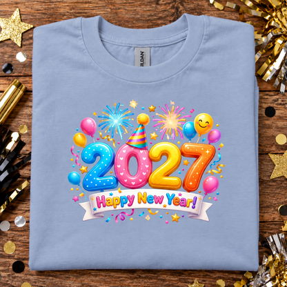 A folded light blue t-shirt with a colorful '2027 Happy New Year!' graphic print, displayed on a wooden surface surrounded by gold and black party decorations.