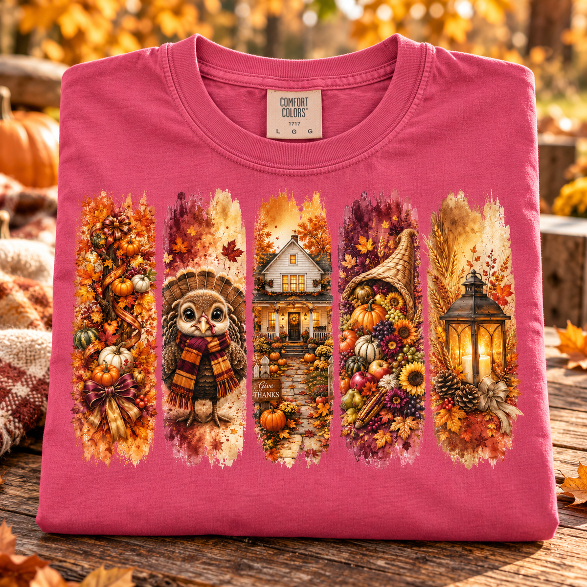A folded crunchberry pink Comfort Colors t-shirt is displayed on a rustic wooden surface with a blurred autumn garden background. The shirt features a large, detailed five-panel graphic print depicting fall harvest scenes, including pumpkins, an owl in a scarf, a house, a cornucopia, and a lantern.