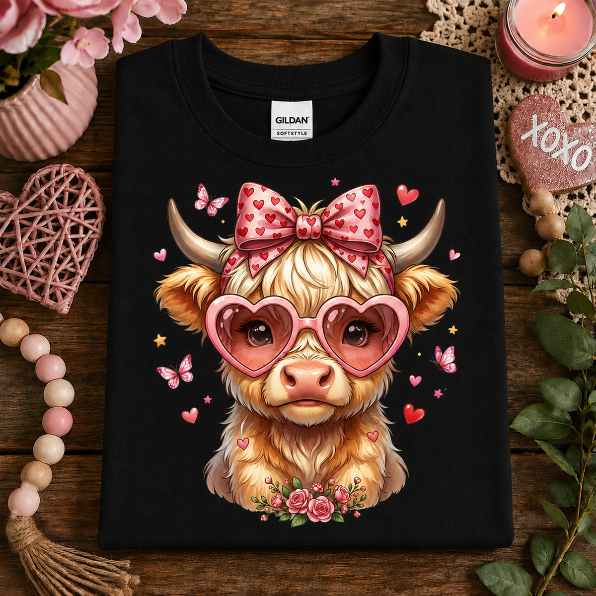 A black Gildan Comfortshorts t-shirt featuring a cute cartoon illustration of a fluffy light-brown cow wearing pink heart-shaped sunglasses and a pink bow with red hearts. The shirt is displayed in a styled flat-lay on a rustic wooden table surrounded by decorative pink and natural elements.
