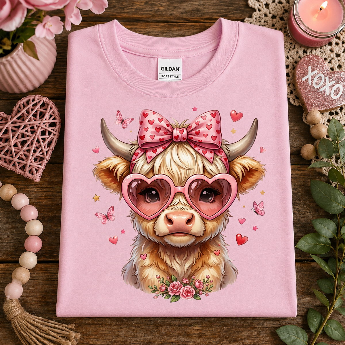 A folded light pink Gildan t-shirt with a cute graphic of a fluffy Highland cow wearing pink heart sunglasses and a pink bow with hearts, styled on a rustic wooden table with Valentine's themed decorations.