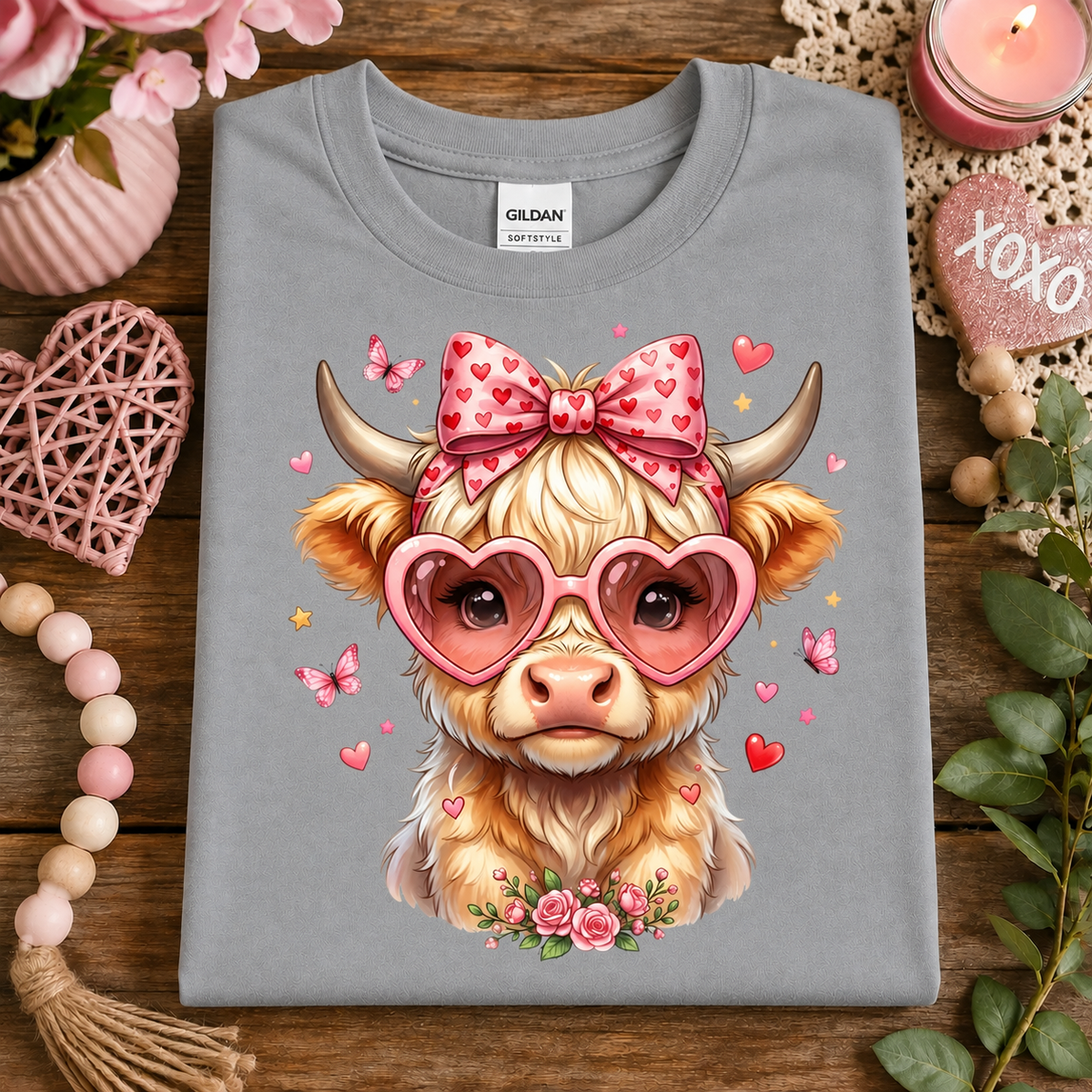 A folded sport gray t-shirt with a cute graphic of a fluffy light brown cow wearing pink heart-shaped glasses and a pink bow with red hearts, styled on a wooden surface with decorative props.