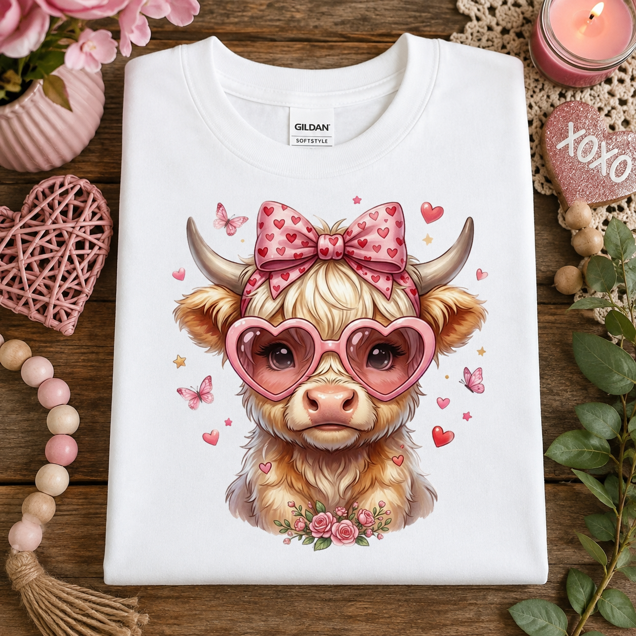 A folded white Gildan Softstyle T-shirt with a colorful graphic of a fluffy light brown cow wearing pink heart-shaped sunglasses and a pink bow with hearts, surrounded by pink hearts, butterflies, and flowers, styled on a wooden surface with Valentine's themed props.