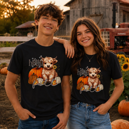 Two people wearing black t-shirts with a highland cow and pumpkin design in a farm setting.
