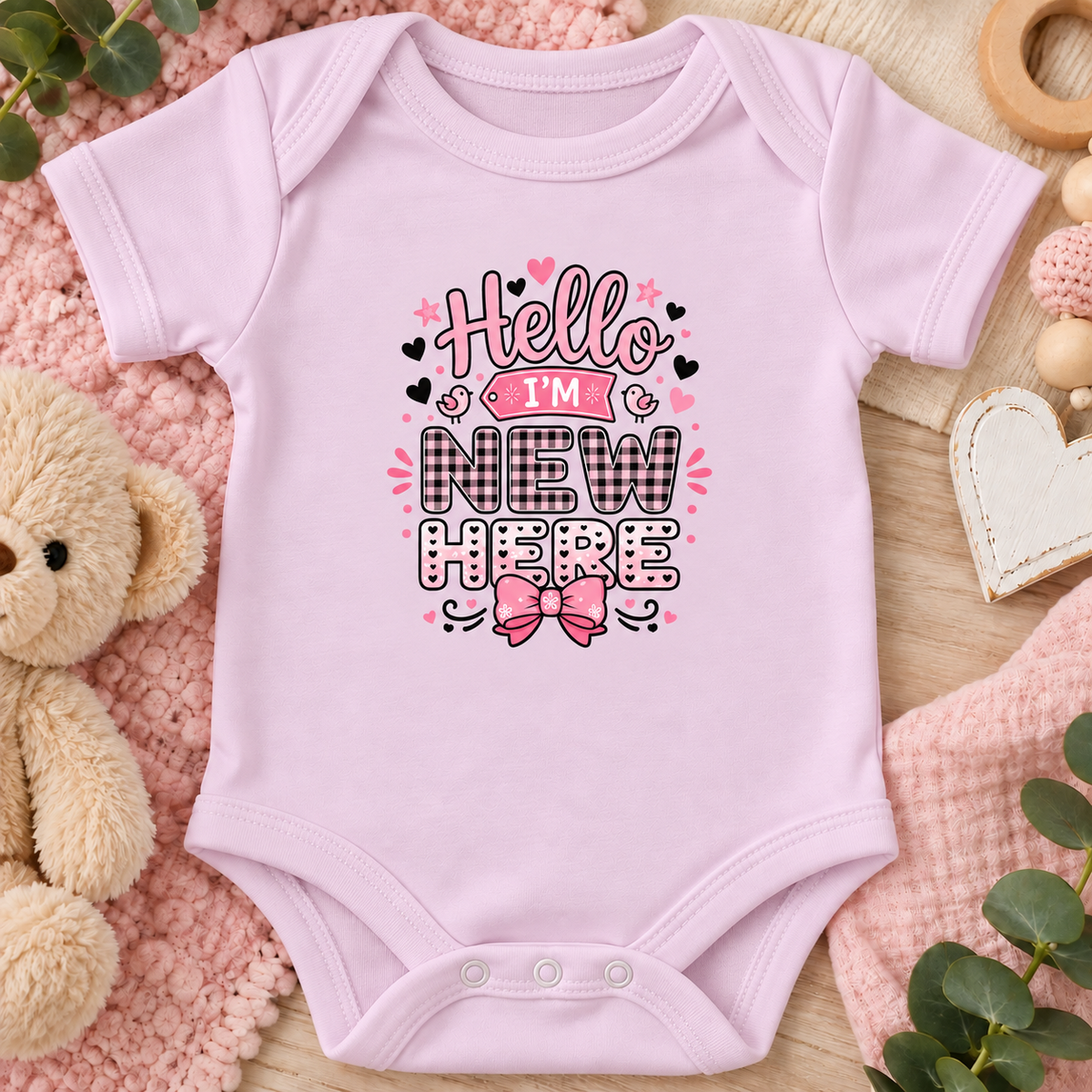 A pink short-sleeve baby bodysuit laid flat on a soft pink textured blanket, surrounded by a teddy bear, wooden beads, a heart-shaped ornament, and green leaves. The bodysuit features a graphic print that reads 'Hello I'M NEW HERE' in black, pink, and white lettering with a pink bow illustration.