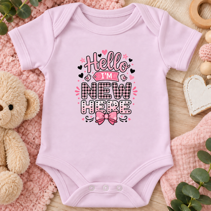 A pink short-sleeve baby bodysuit laid flat on a soft pink textured blanket, surrounded by a teddy bear, wooden beads, a heart-shaped ornament, and green leaves. The bodysuit features a graphic print that reads 'Hello I'M NEW HERE' in black, pink, and white lettering with a pink bow illustration.