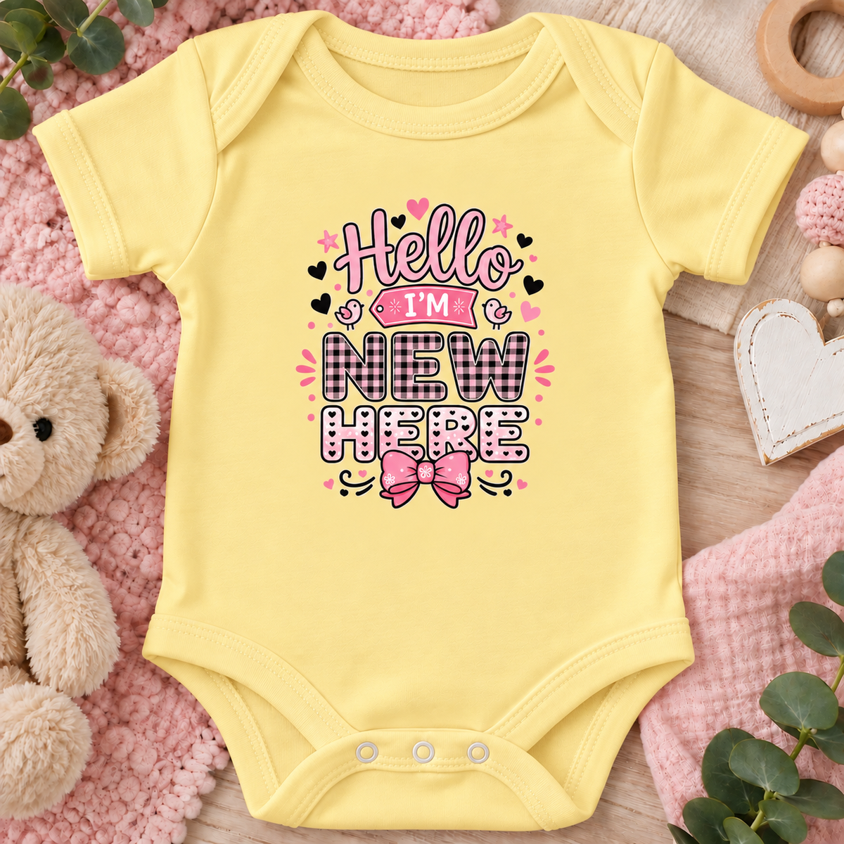 A butter yellow short-sleeve baby bodysuit laid flat on a pink textured blanket with a teddy bear, wooden beads, and greenery around it. The front has a decorative printed phrase reading “Hello I’m New Here” in pink, black, and white with a pink bow graphic.