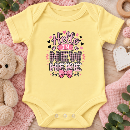 A butter yellow short-sleeve baby bodysuit laid flat on a pink textured blanket with a teddy bear, wooden beads, and greenery around it. The front has a decorative printed phrase reading “Hello I’m New Here” in pink, black, and white with a pink bow graphic.